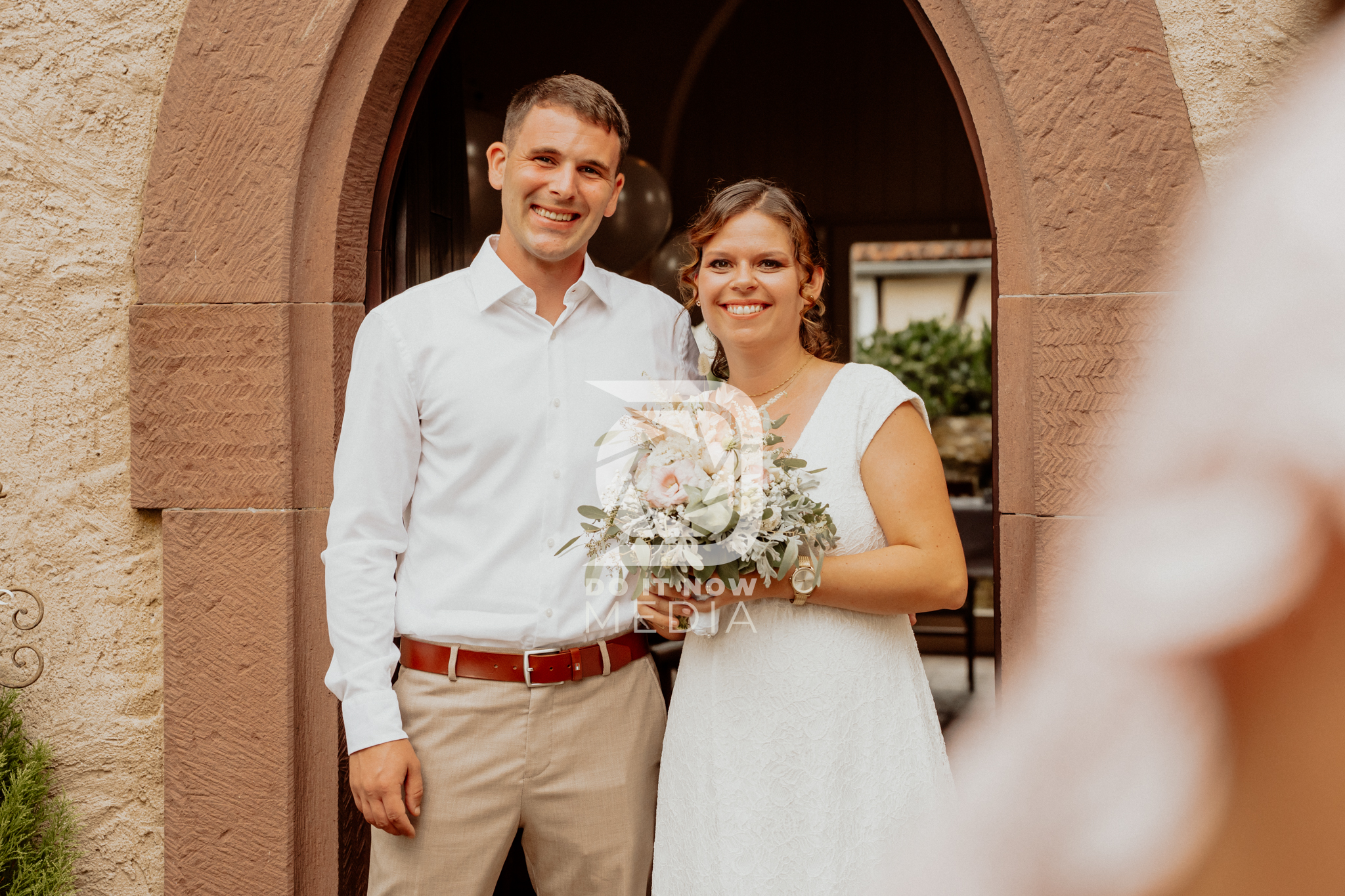 Jessica und Julian lächeln gemeinsam im steinernen Torbogen, Jessica hält ihren Brautstrauß.